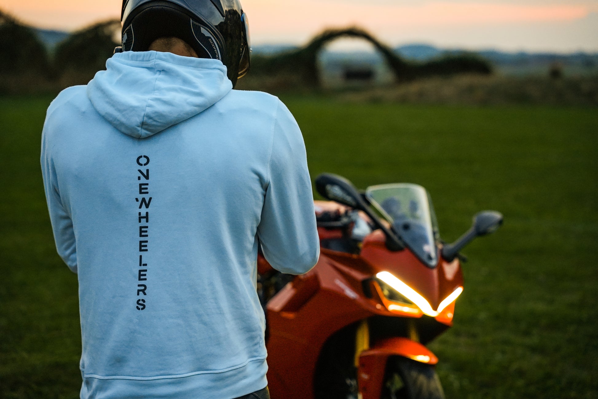 Back view of a rider wearing the white Onewheelers Crew Hoodie next to a Ducati 950 at sunset, showcasing premium Onewheelers motorcycle streetwear.